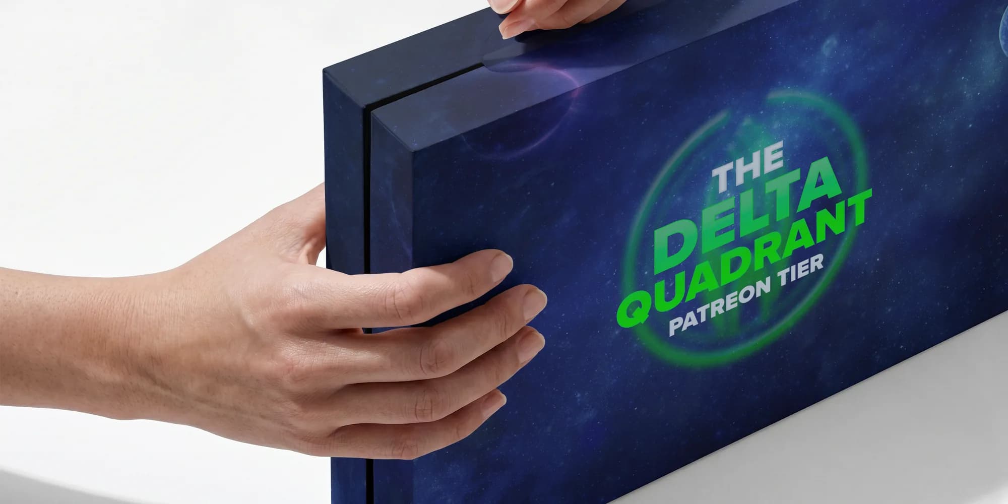 A Delta Quadrant Patreon tier box in deep space blue and green, held in two hands