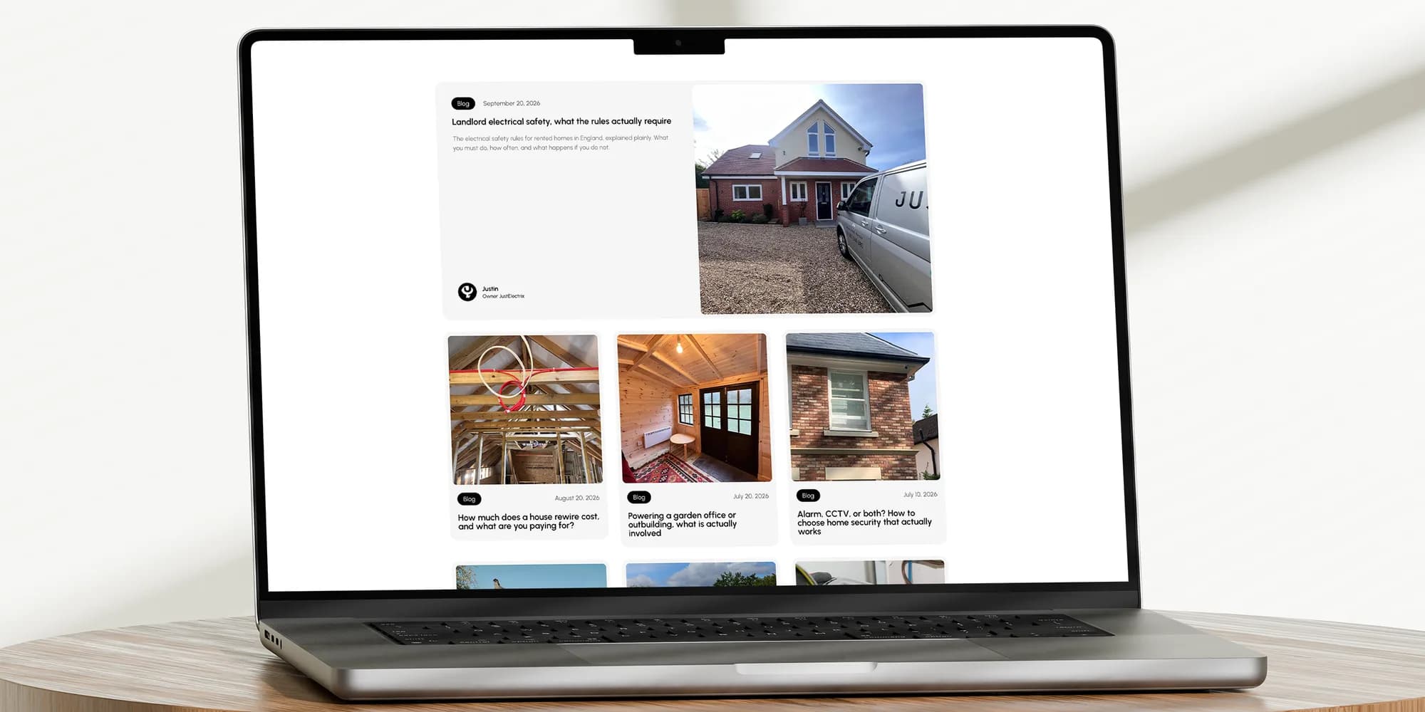 The JustElectrix blog on a MacBook Pro on a round wooden table: landlord safety, rewire costs, garden offices, home security